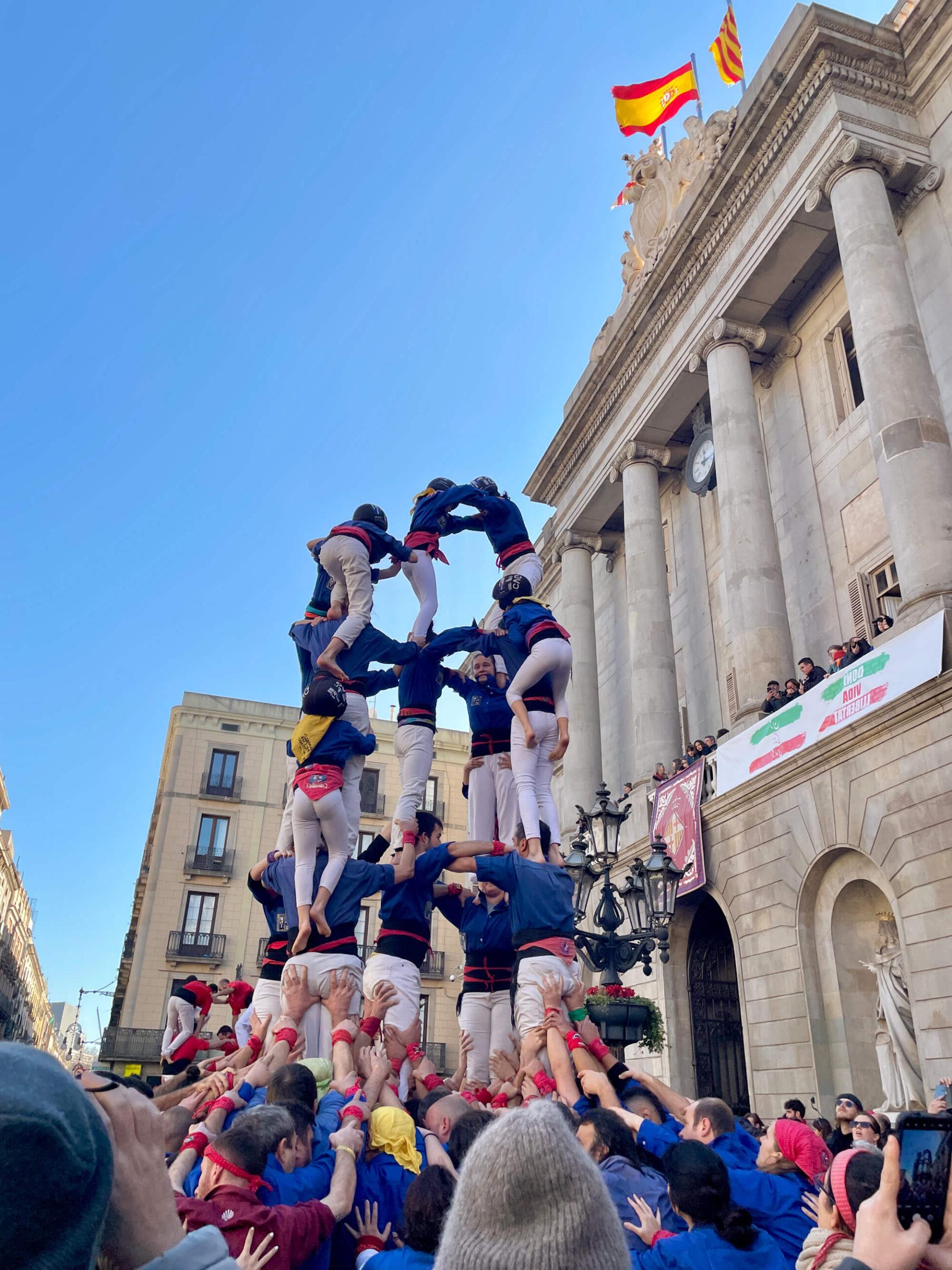 A human tower is formed by a group of people, standing in front of a historic building with columns. 在楼顶可以看到旗帜. 塔底很宽, 到达顶部时逐渐变窄, 有几个层次的参与者. 围观者聚集在周围.