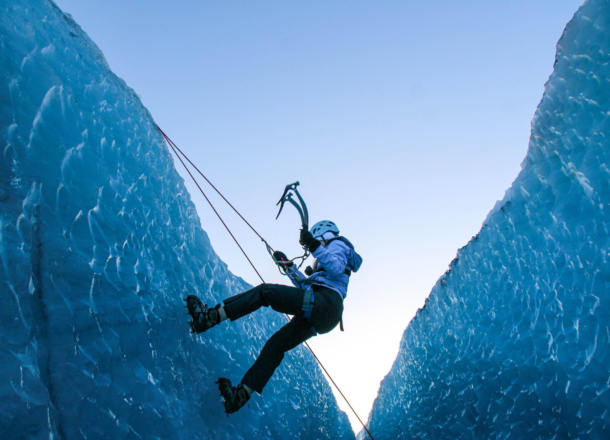 攀岩者:穿着攀岩装备的人, 包括头盔和冰爪, 冰在两堵高高的冰墙之间攀爬吗. 他们使用冰镐并用绳索固定. 背景中的天空清澈明亮.