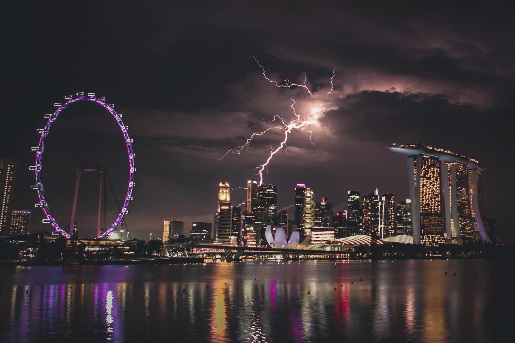 一个充满活力的夜间新加坡城市景观与闪电击中天空. 滨海湾金沙酒店的天际线灯火通明, 莲花形的艺术科学博物馆, 还有闪闪发光的新加坡摩天轮, 一切都映在宁静的水面上.