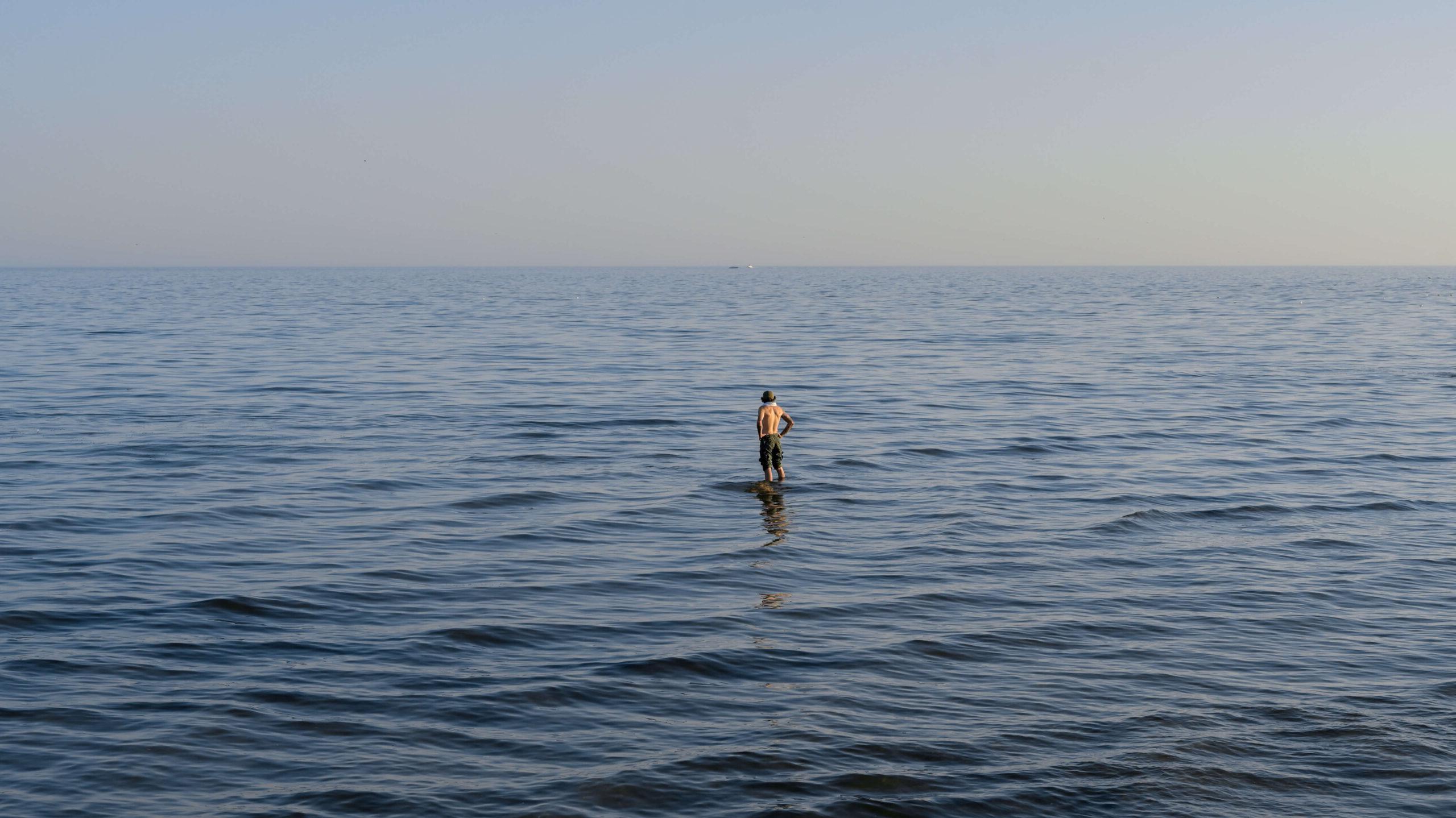 A person stands in the expansive, calm sea under a clear sky, with water stretching to the horizon. Their reflection is visible on the water's surface, creating a tranquil and serene scene.