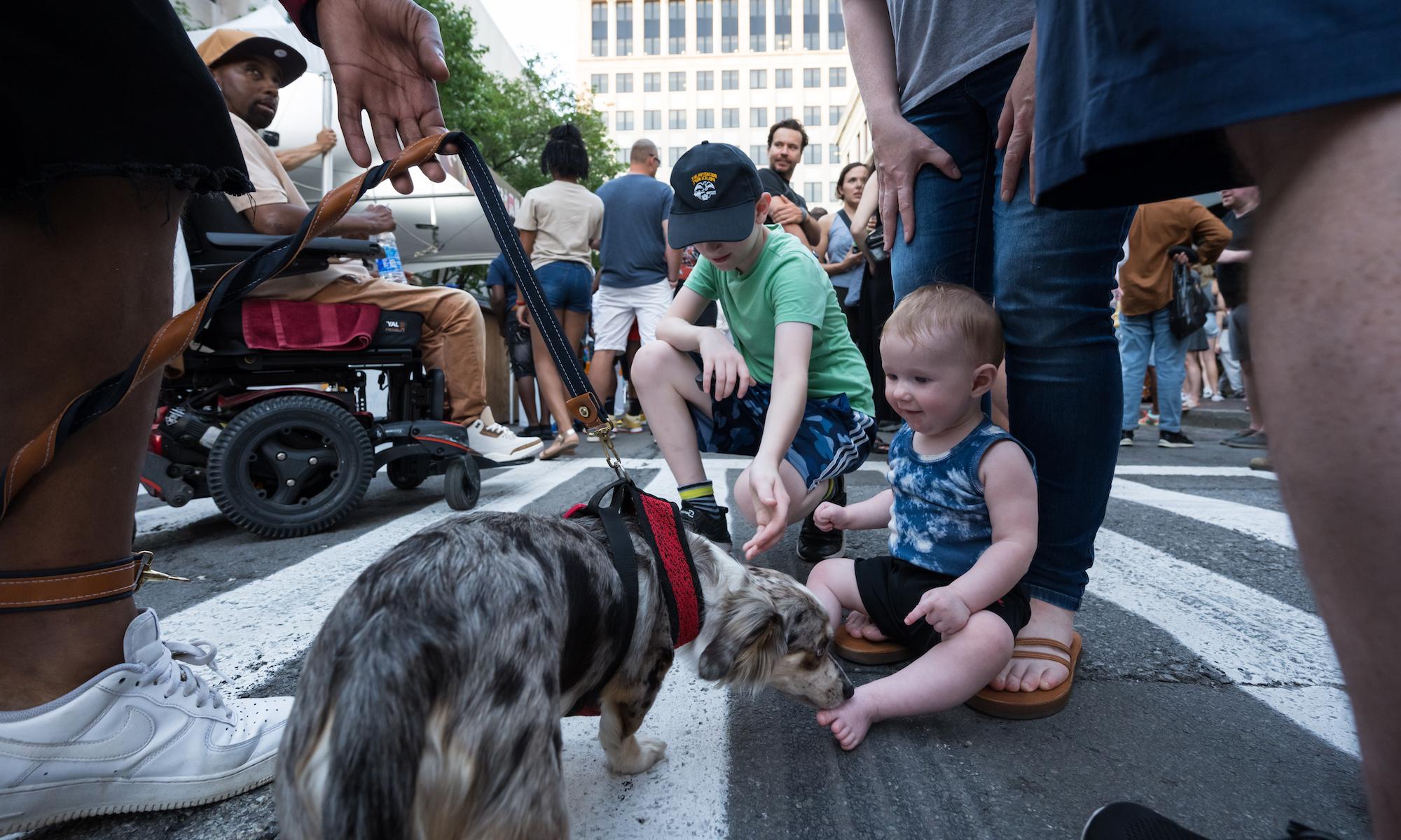 一个婴儿和一个小孩在外面忙碌的人群中抚摸着一只狗